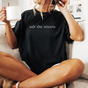 May include: Black t-shirt with the text "salt the streets" in white. The shirt is a casual style, with a relaxed fit. A white mug is held in the left hand. The person is sitting on a couch.