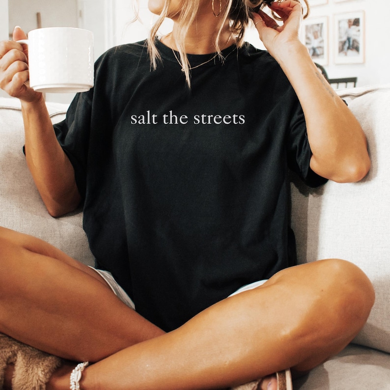 May include: Black t-shirt with the text "salt the streets" in white. The shirt is a casual style, with a relaxed fit. A white mug is held in the left hand. The person is sitting on a couch.