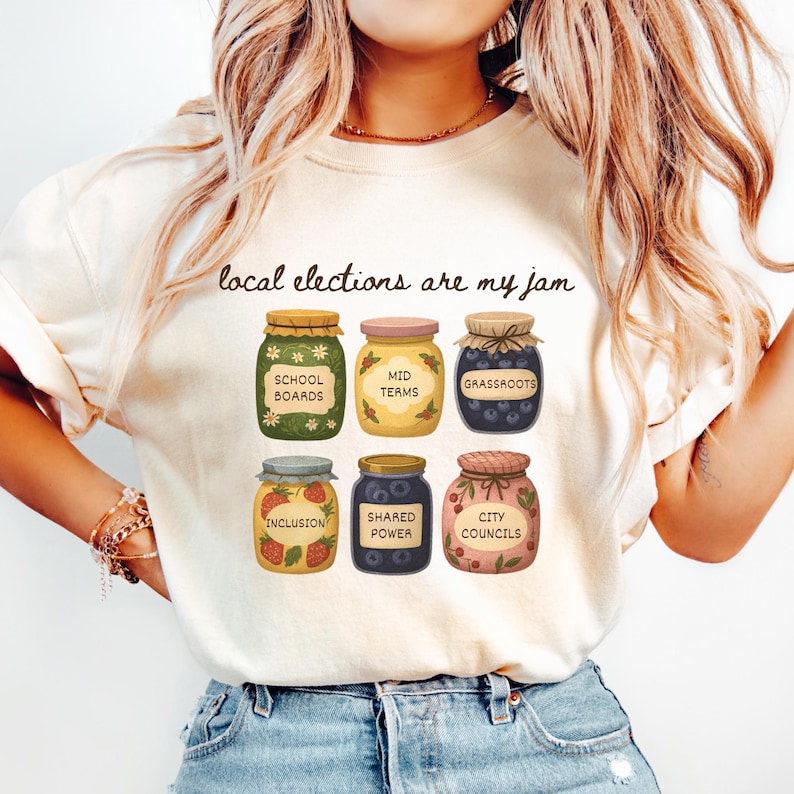 May include: Cream-colored t-shirt with the phrase "local elections are my jam" above illustrations of six jam jars. Each jar has a different label: "School Boards," "Mid Terms," "Grassroots," "Inclusion," "Shared Power," and "City Councils."