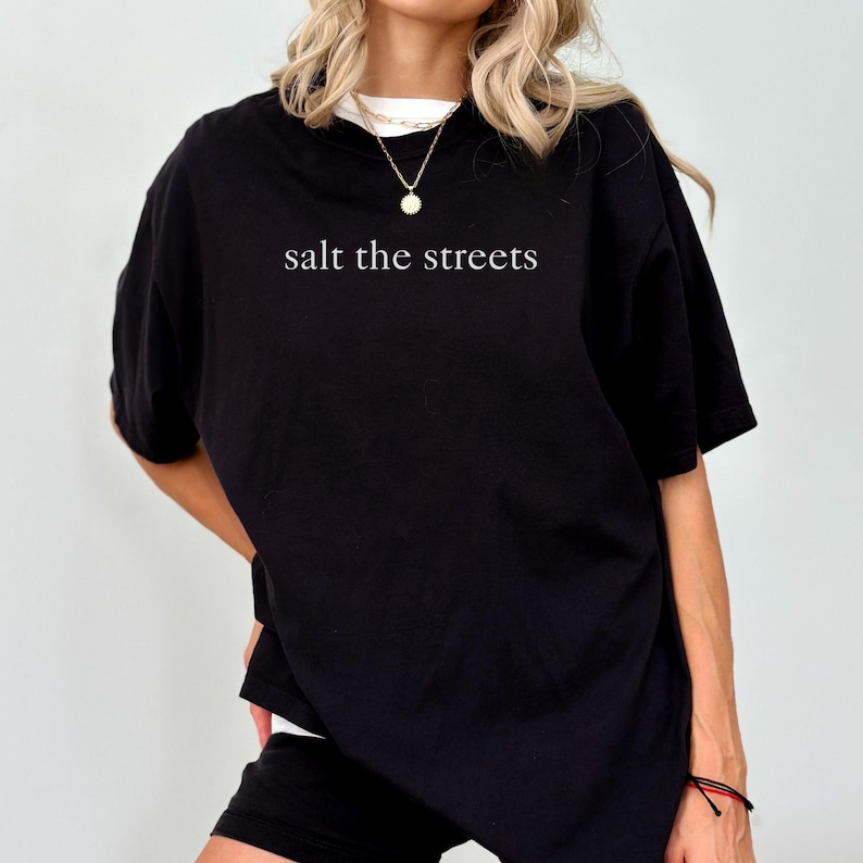 May include: Black oversized t-shirt with the phrase "salt the streets" printed in white. The shirt has short sleeves and a crew neck. The model is wearing a gold necklace.