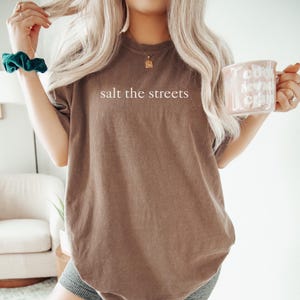 May include: A brown t-shirt with the text "salt the streets" in white. The shirt is worn by a person with long blonde hair. A teal scrunchie is on the wrist. A pink mug with white text is held in the hand.
