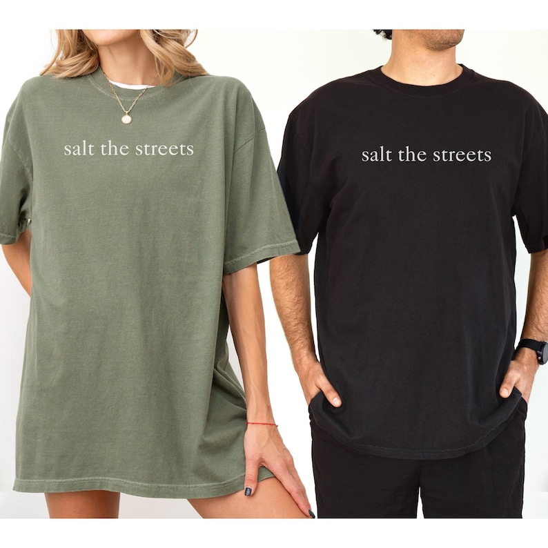 May include: Two short-sleeved t-shirts, one olive green and one black, are displayed. The phrase "salt the streets" is printed in white across the chest of each shirt. The shirts appear to be made of a soft, comfortable material.