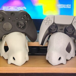 May include: Two video game controllers, one black and one white, are displayed on a white skull-shaped stand. The controllers are resting on the stand, with the black controller on the left and the white controller on the right.