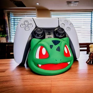 May include: A white PlayStation 5 controller with a green Bulbasaur character faceplate. The controller is sitting on a wooden table.