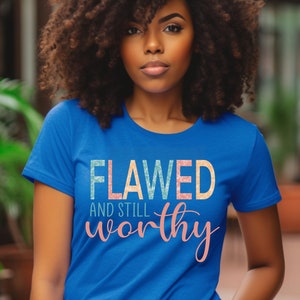 May include: A royal blue t-shirt with the text "Flawed and still worthy" in a colorful, glittery font.