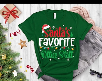 May include: A green t-shirt with the text "Santa's Favorite Office Staff" in red, white, and green. The design includes a Santa hat, Christmas lights, and snowflakes. The shirt is folded and tied at the bottom, with a pair of jeans and accessories in the background.