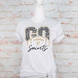 May include: A gray t-shirt with a gold football design featuring the number "90" in a black and white leopard print. The text "Saints" is written in gold cursive script below the football.