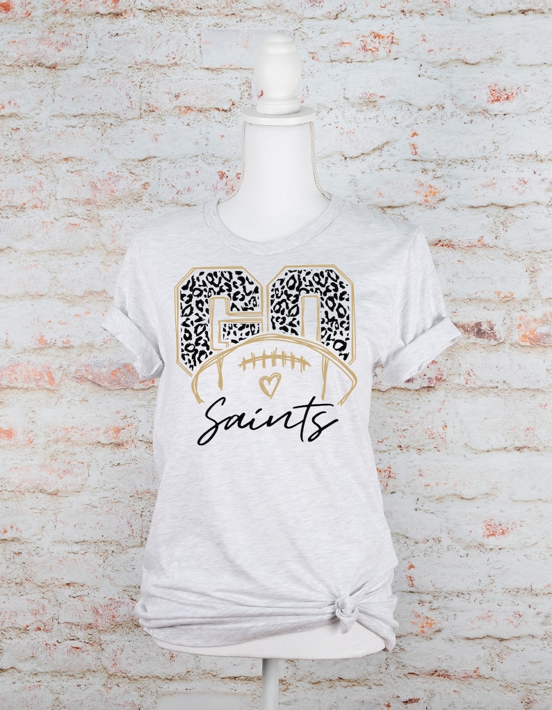 May include: A gray t-shirt with a gold football design featuring the number "90" in a black and white leopard print. The text "Saints" is written in gold cursive script below the football.