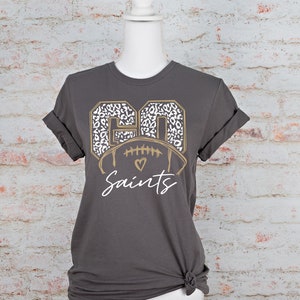 May include: A gray t-shirt with a gold and white football graphic. The football is surrounded by the numbers "60" in a leopard print. The text "Saints" is below the football.