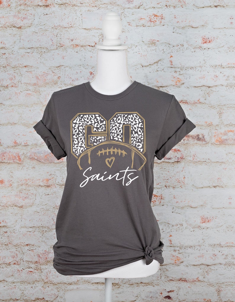 May include: A gray t-shirt with a gold and white football graphic. The football is surrounded by the numbers "60" in a leopard print. The text "Saints" is below the football.