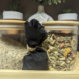 Puede incluir: Dos tarros de vidrio con tapas de madera, uno lleno de hierbas y flores secas, el otro con una sustancia marrón claro. Una pequeña bolsa de tela negra atada con una cuerda se encuentra entre los tarros.