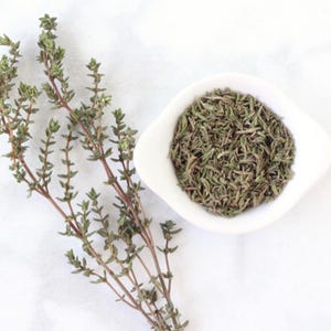 May include: Fresh thyme sprigs and dried thyme in a white bowl. The thyme is a green herb with a strong aroma.