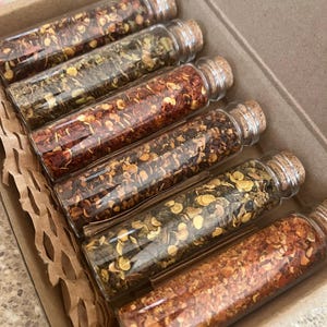 May include: Six small glass jars with cork tops filled with different types of dried red pepper flakes and herbs. The jars are arranged in a row in a brown cardboard box with brown paper packing material.