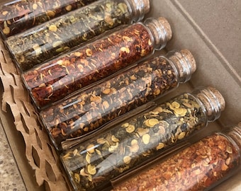 May include: Six small glass jars with cork tops filled with different types of dried red pepper flakes and herbs. The jars are arranged in a row in a brown cardboard box with brown paper packing material.