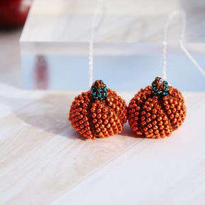 Peut inclure: Deux boucles d'oreilles en forme de citrouille perlées. Les boucles d'oreilles sont orange avec une surface texturée et une petite tige perlée verte et marron. Elles sont suspendues à un fin fil transparent.