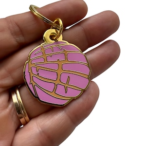 May include: A gold-colored keychain with a pink and gold enamel charm shaped like a concha, a traditional Mexican sweet bread.