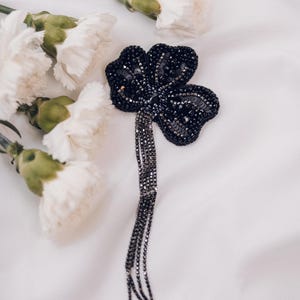 May include: A black, four-leaf clover brooch, heavily embellished with sequins and beads, with dangling chain tassels. The brooch is set against a white background with white carnation flowers.