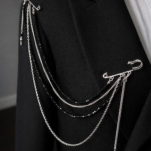 Puede incluir: Broche de hombro plateado con múltiples cadenas y cuentas negras, sujeto con imperdibles a una blazer negra. Un pequeño dije de estrella cuelga de una cadena. El broche añade un acento único y atrevido a la prenda.