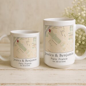 May include: Two white ceramic mugs with a map design of Paris, France. Each mug features the names "Jessica & Benjamin" and the date "Est. 06/27/2025". A red pin marks a location on the map. One mug is larger than the other.