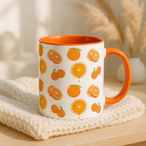 May include: White ceramic mug with an orange interior and handle. The mug is decorated with a pattern of oranges and orange slices. The mug is sitting on a cream-colored knitted coaster.