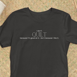 May include: Dark gray t-shirt with the text "I don't QUILT because I'm good at it. I do it because I like it." The shirt is displayed on a woven mat with pears and pottery tools.