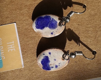 May include: A pair of white ceramic earrings with blue accents. The earrings are oval shaped and have silver hooks. The earrings are displayed on a brown card with the text "The Gilding Experience".