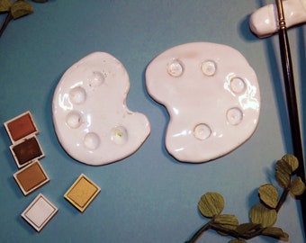 May include: Two white ceramic paint palettes with round wells for mixing paint. The palettes are on a blue surface with watercolor paint pans and a small branch with green leaves.