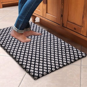 May include: A rectangular black and white patterned kitchen mat. The mat features a repeating diamond design. The mat is placed on a tiled floor, with a person's feet standing on it. Wooden cabinets are in the background.