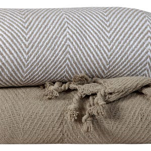 May include: Two folded blankets with a herringbone pattern. The top blanket is white and tan, while the bottom blanket is solid tan with tassels. The blankets are made of a soft, woven material.