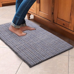 May include: A rectangular, blue and gray woven mat with a textured surface. The mat is placed on a tiled floor, with a person's feet and legs visible. The mat is positioned in front of a wooden cabinet.