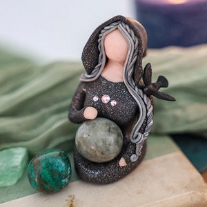 The Crone is dressed in a sparkling black robe and has long silver hair. She has three rhinestones across her chest . She has the raven or crow on her shoulder, the messenger between worlds, and she holds a labradorite crystal in her lap.