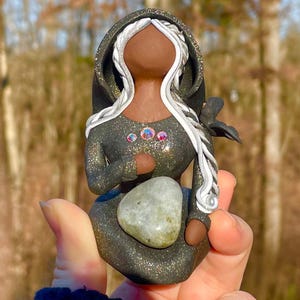 May include: A handmade figurine of a woman with long white hair, wearing a black hooded cloak with silver accents and holding a grey stone. The figurine is decorated with iridescent gems and has a black crow perched on her shoulder.