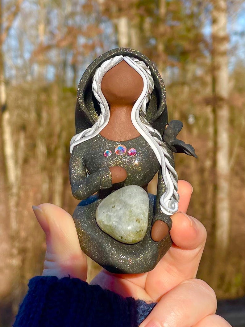 May include: A handmade figurine of a woman with long white hair, wearing a black hooded cloak with silver accents and holding a grey stone. The figurine is decorated with iridescent gems and has a black crow perched on her shoulder.