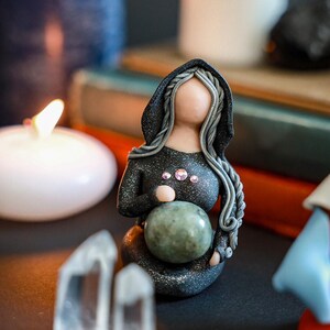 May include: A black and silver figurine of a woman wearing a hooded cloak, holding a green sphere. The figurine is surrounded by candles and crystals.