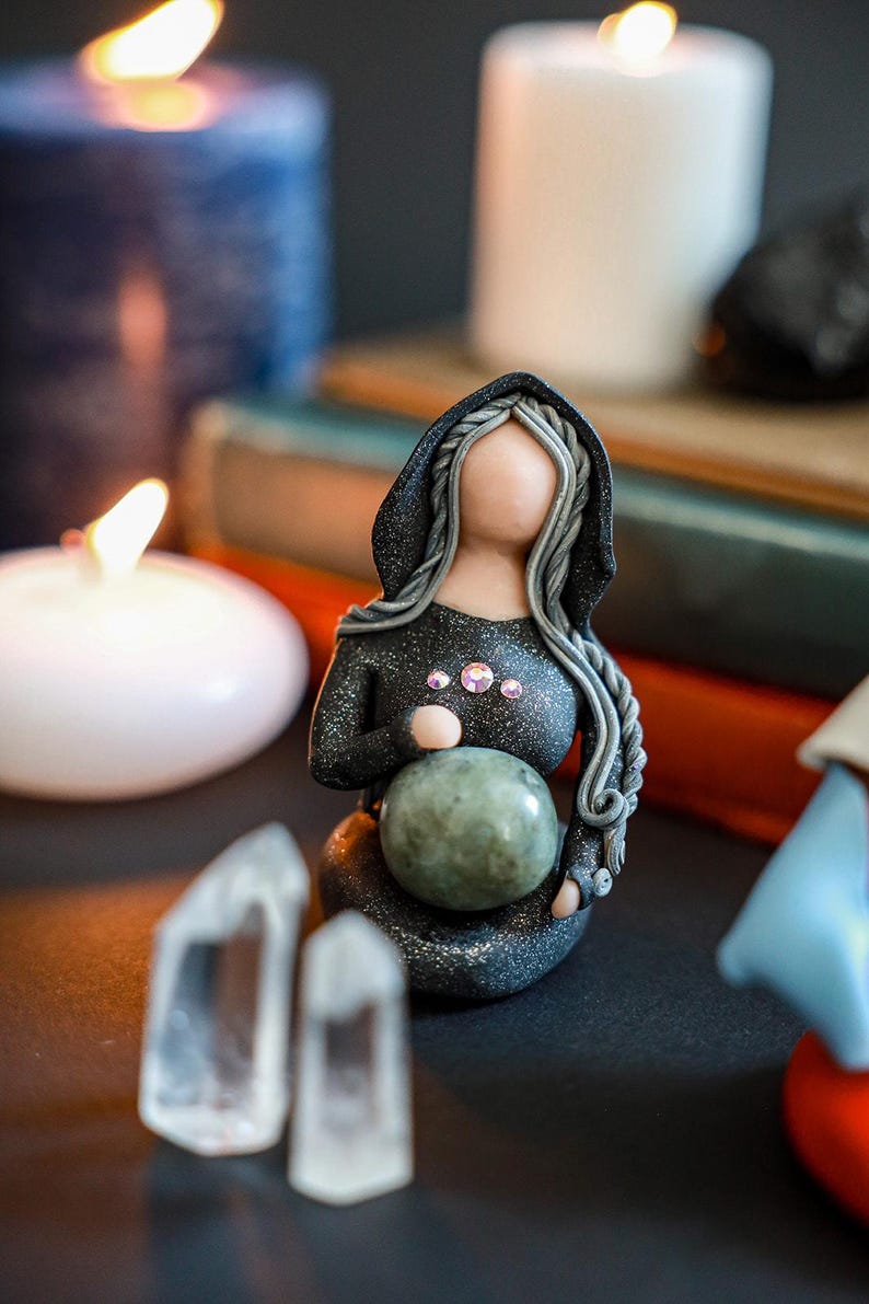 May include: A black and silver figurine of a woman wearing a hooded cloak, holding a green sphere. The figurine is surrounded by candles and crystals.