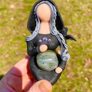May include: A black and silver figurine of a woman with long hair, wearing a hooded cloak and holding a green stone. She has a small bird perched on her shoulder.