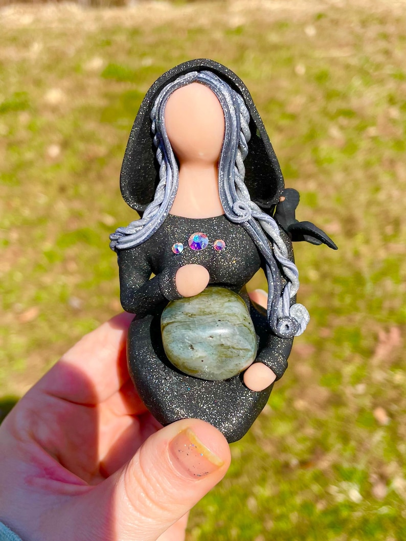 May include: A black and silver figurine of a woman with long hair, wearing a hooded cloak and holding a green stone. She has a small bird perched on her shoulder.