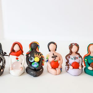 May include: Seven small, colorful, handmade figurines of women, each holding a different object. The figurines are made of clay and have painted details. They are all sitting in a row on a white surface.