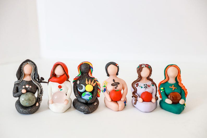 May include: Seven small, colorful, handmade figurines of women, each holding a different object. The figurines are made of clay and have painted details. They are all sitting in a row on a white surface.