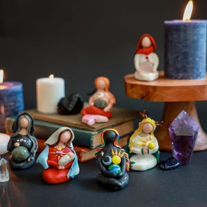 May include: A collection of small, colorful figurines of women holding various objects, including a baby, a crystal, and a globe. The figurines are arranged on a table with candles and crystals.