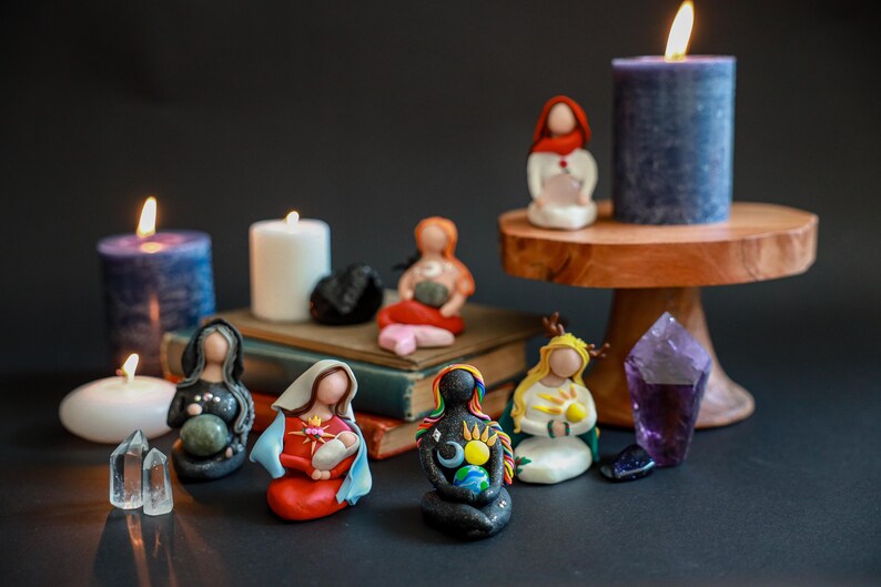 May include: A collection of small, colorful figurines of women holding various objects, including a baby, a crystal, and a globe. The figurines are arranged on a table with candles and crystals.