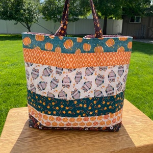 May include: A large tote bag with a patchwork design featuring owls, pumpkins, and fall leaves. The bag has brown straps and is made of a variety of fabrics in orange, green, and white.