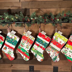 May include: Six Christmas stockings with various patterns, including stripes, holiday characters, and text. The stockings are red, green, and white, and are hanging from a black rod with a garland of greenery.