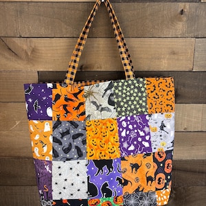 May include: A quilted tote bag featuring a patchwork design of various Halloween-themed fabrics, including black cats, pumpkins, ghosts, skeletons, bats, and spiderwebs in orange, purple, black, and white, with two orange and black plaid fabric handles.