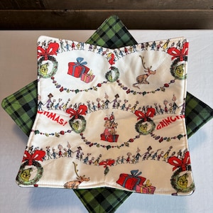 May include: A Christmas-themed bowl cozy with a white background featuring a repeating pattern of characters, gifts, and wreaths. The cozy is layered over a green and black plaid square. The word "Christmas" is visible.
