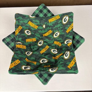 May include: Green Bay Packers themed bowl cozy. The cozy is star-shaped with a green and black plaid pattern on the outside and a green camouflage pattern on the inside. The inside pattern features the Packers logo and the word "PACKERS" in yellow.