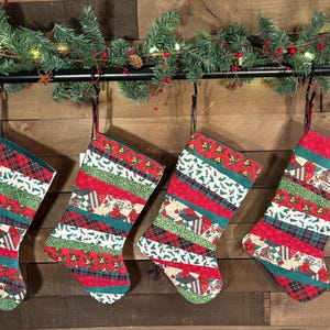 May include: Four quilted Christmas stockings with red, green, and white patchwork designs. Each stocking is suspended from a black rod with a garland of greenery and red berries. The stockings have a variety of patterns, including plaid and floral motifs.