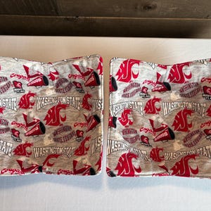 May include: Two gray and red fabric bowl cozies. The cozies are patterned with the words "Washington State" and "Cougars" along with the school's logo. The cozies are square-shaped and designed to hold bowls.