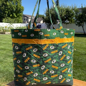 May include: A green and yellow tote bag featuring the Packers logo and text. The bag has a quilted design with a camouflage pattern and a yellow stripe. The handles are green and the bag is sitting on a wooden surface.