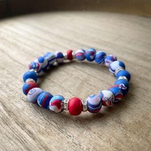 May include: A red, white, and blue patterned bracelet with silver accents. The bracelet is made of round beads and has a single red bead in the center.